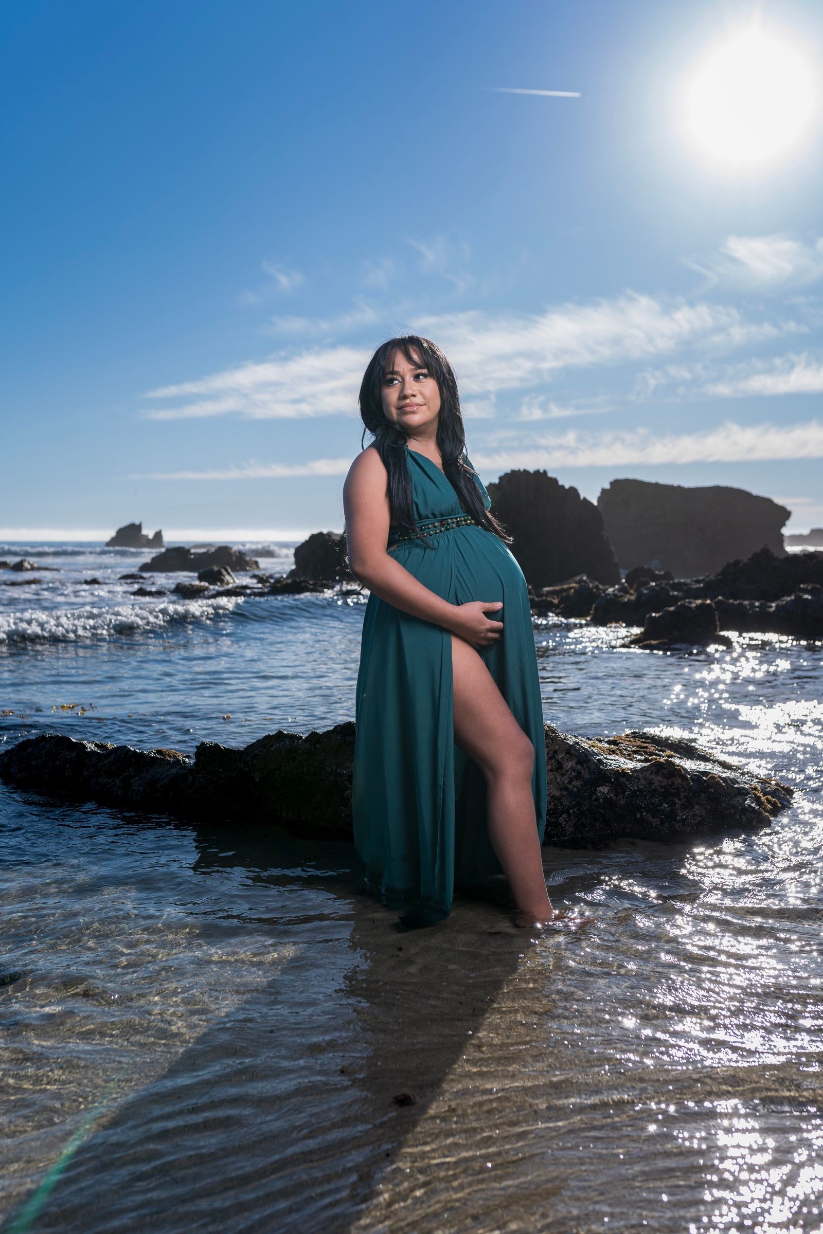 Expectant mother in a green gown standing beside the rocky coast.