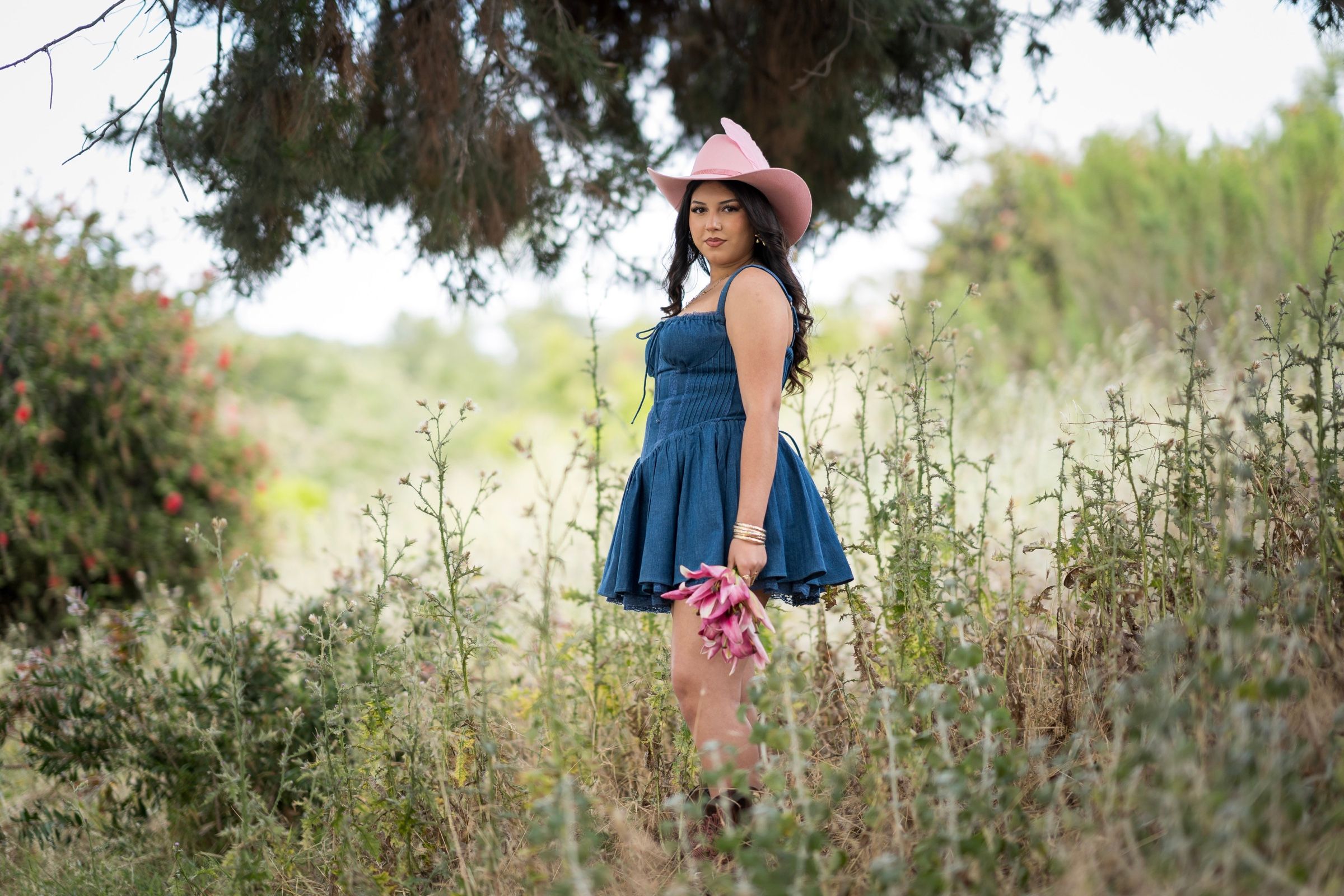 Young woman in a blue dress and pink cowboy hat holding flowers outdoors.