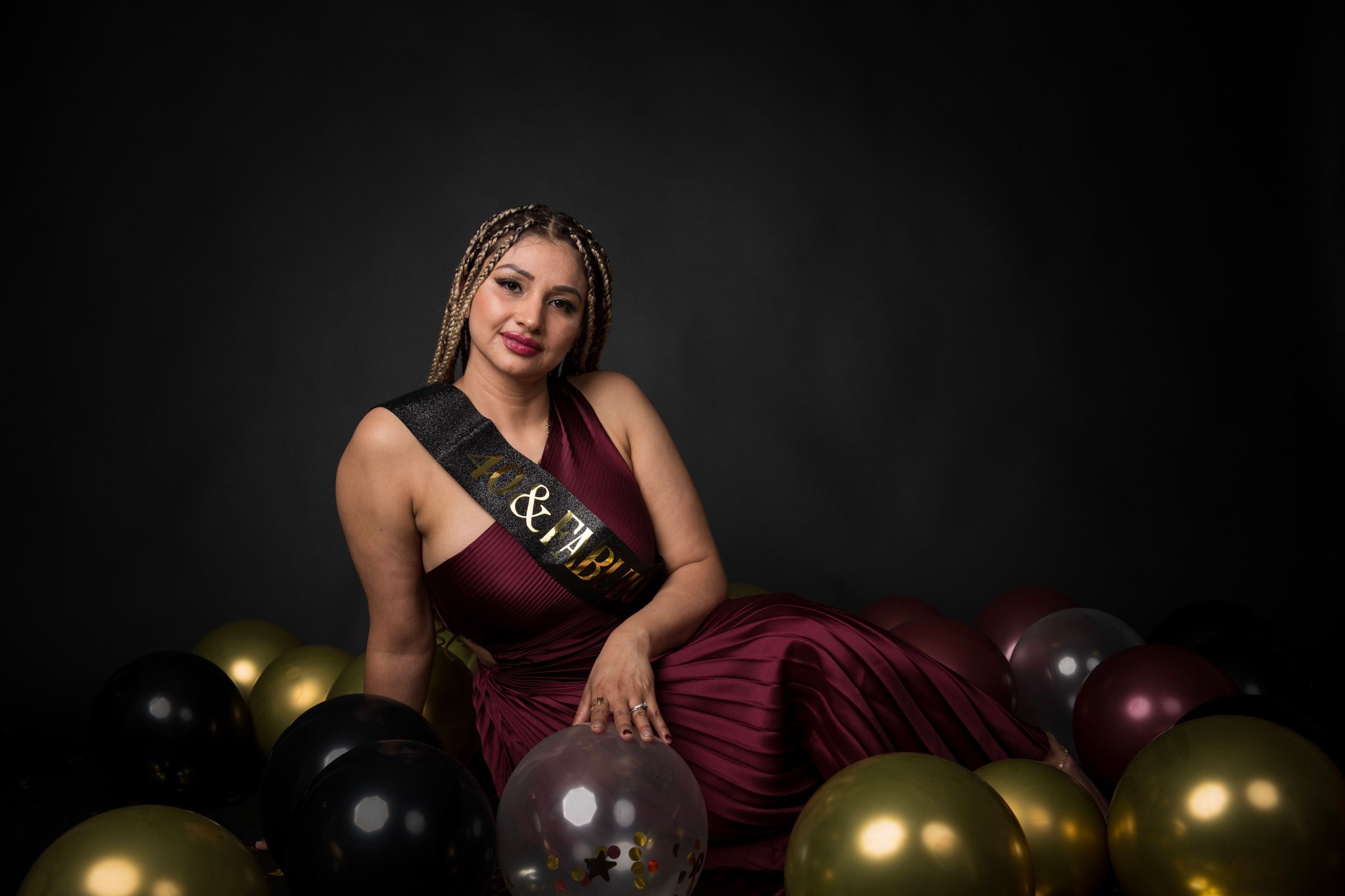 Woman in a burgundy gown posing among black and gold birthday balloons.