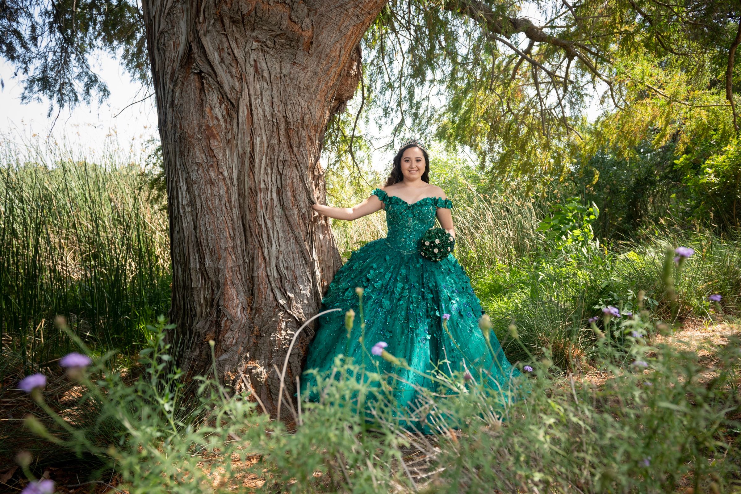 Quinceañera in an emerald gown posing beside a large tree outdoors.