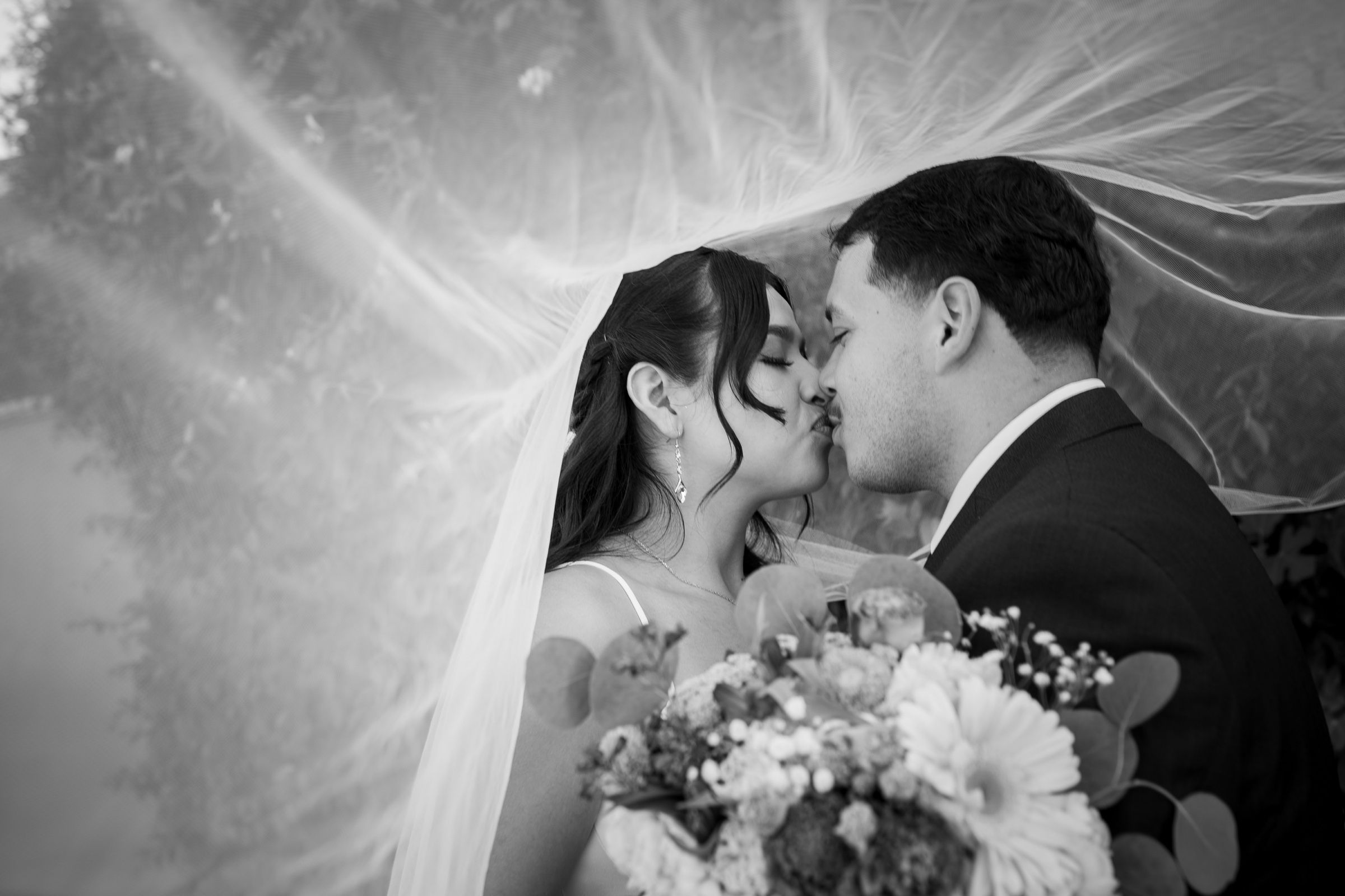 Newlyweds kissing beneath a flowing veil in a black-and-white portrait.